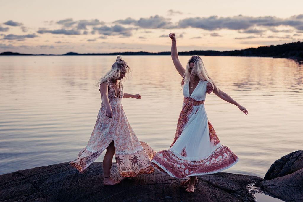Två kvinnor dansar i långa klänningar på en klippa vid havet i skymningen.