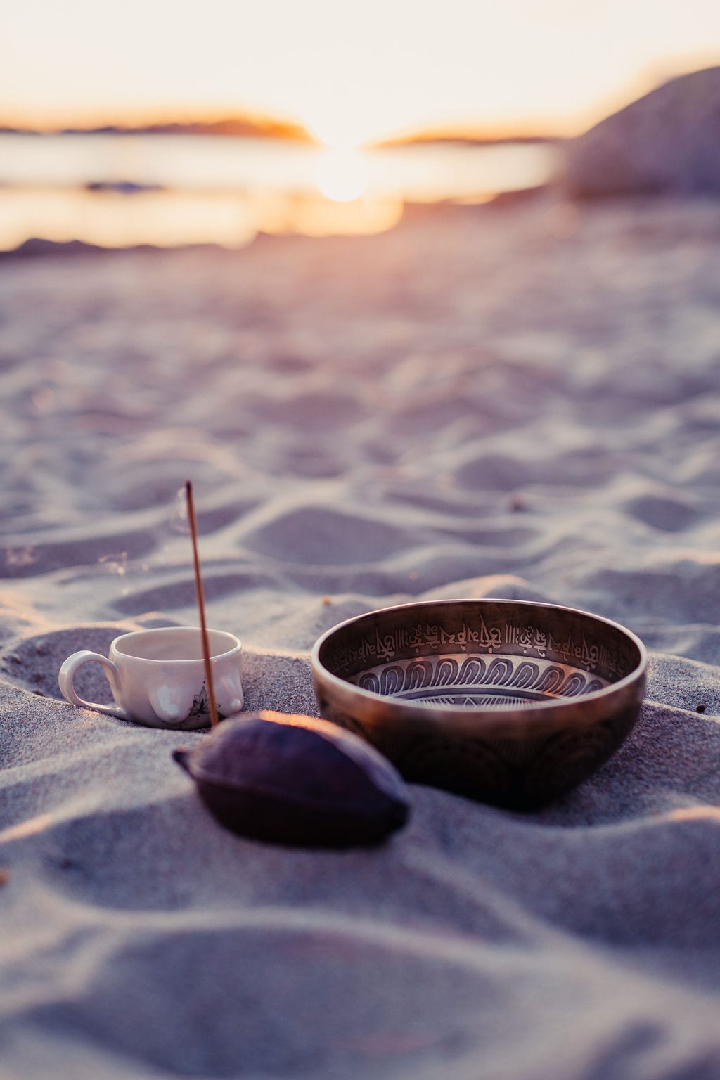 Rökelsepinne, klangskål, kopp och kakaofrukt i sanden på en strand.