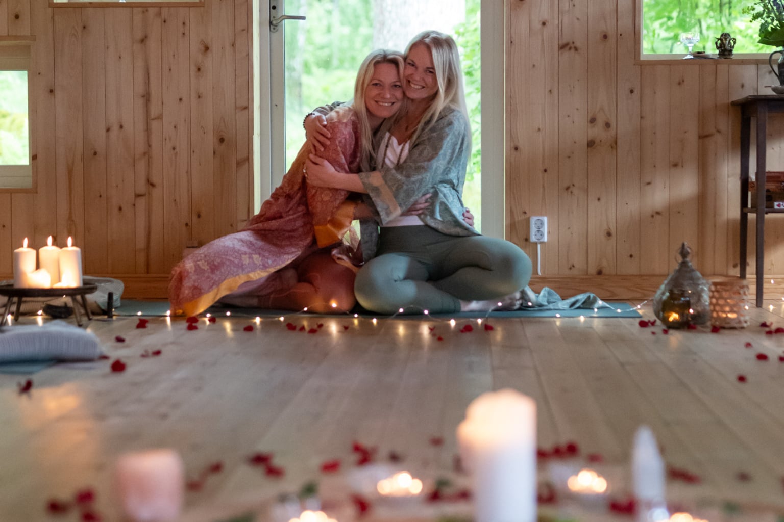 Två kvinnor kramar om varandra på golvet i ett trärum med tända ljus och rosenblad.