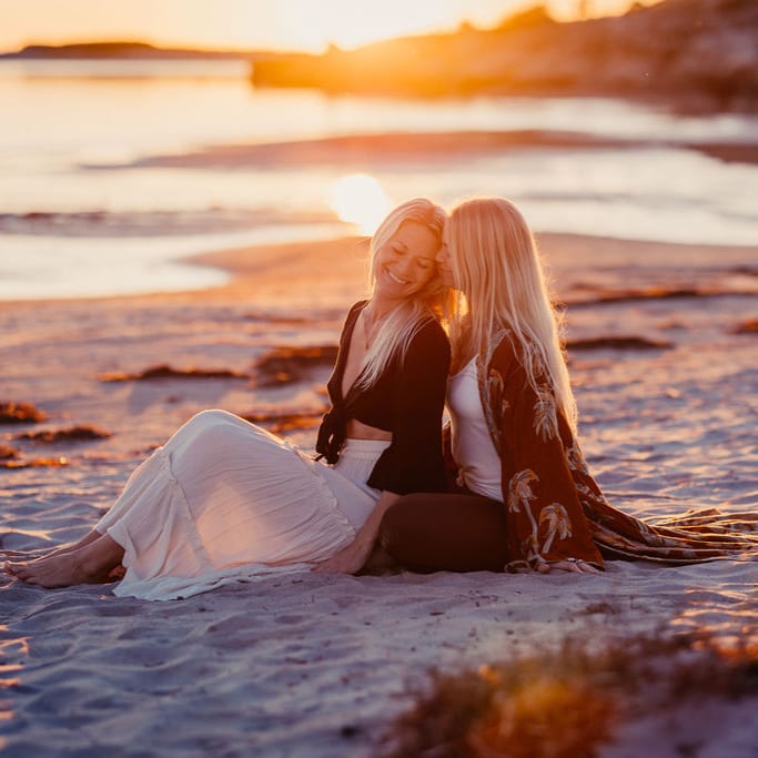 Två kvinnor sitter tätt tillsammans på en sandstrand i solnedgången.