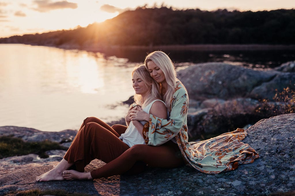 Två kvinnor sitter tätt tillsammans på en klippa vid vattnet i solnedgången.