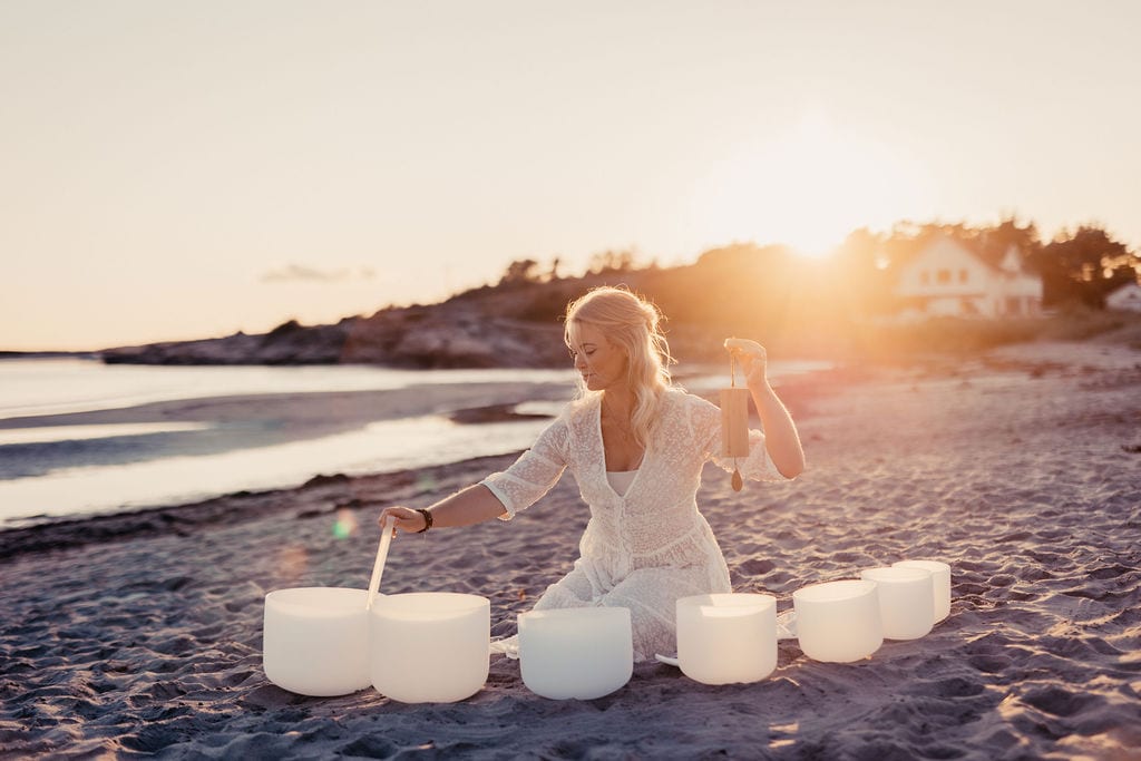 Kvinna spelar på vita kristallskålar på en strand i kvällssol.